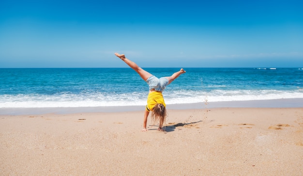 Photo young happy athletic girl in a yellow t-shirt standing on hands on the sand, sea horizon and white waves on a sunny day, vacation freedom joyful movement