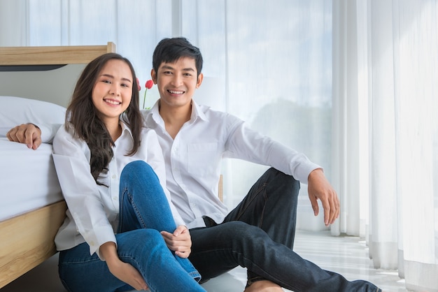 Giovani coppie asiatiche attraenti felici che portano camicia bianca pulita che si siedono insieme sul pavimento in camera da letto che sorride alla macchina fotografica con il fondo bianco della tenda. concetto per amore e relazione felice.