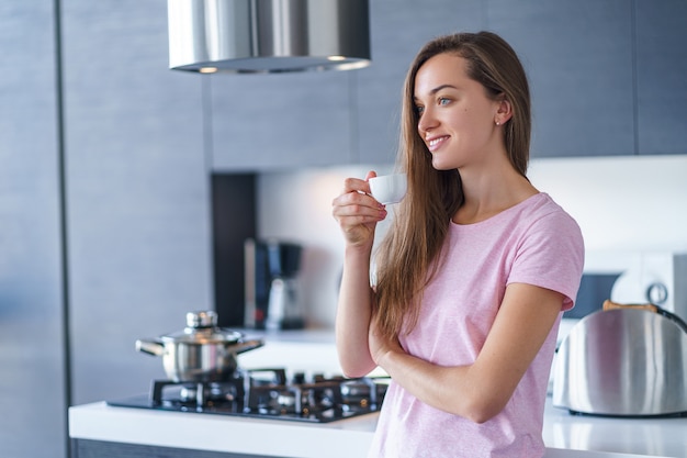 若い幸せな魅力的な白人女性が飲んで、朝早くキッチンで新鮮な香り豊かなコーヒーを自宅で楽しんで