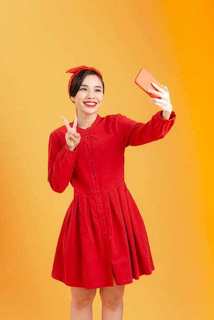 赤いドレスを着た若い幸せな魅力的な女性は、彼女の携帯電話を使用して写真を撮り、カラフルなオレンジ色の背景を撮影します。