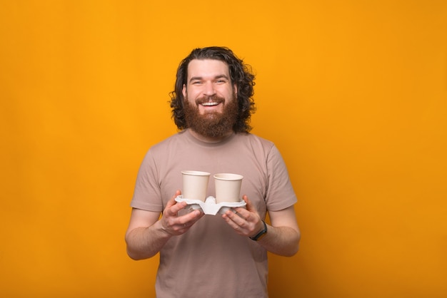 Giovane uomo barbuto felice con i capelli lunghi che tiene due tazze di caffè su giallo