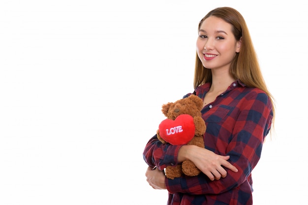 Young happy beautiful Asian woman smiling and holding teddy bear with heart and love sign