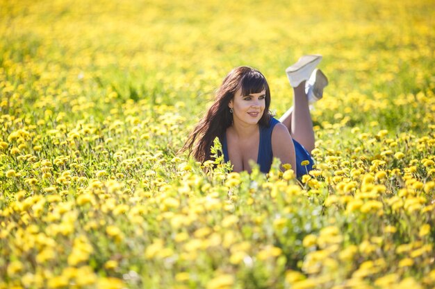 Young happy beautiful big size model girl in blue dress lies resting on blooming dandelions field in summer