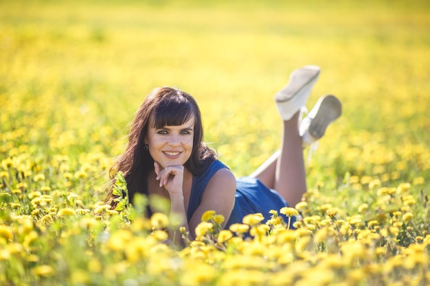 Молодая счастливая красивая девушка большого размера в синем платье лежит летом на цветущем поле одуванчиков