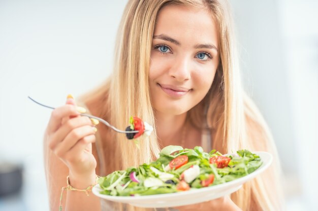 Giovane ragazza bionda felice che mangia insalata sana da rucola spinaci pomodori olive cipolla e olio d'oliva.