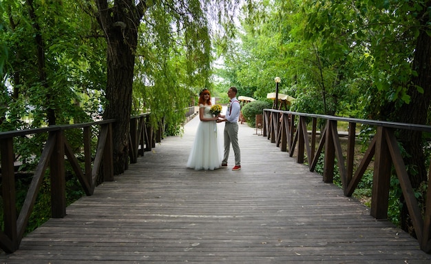 Молодая счастливая невеста и жених держат друг друга за руки и смотрят друг на друга на мосту под зеленой листьями.