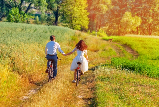 Young happy bride and groom ride bicycles in the meadow back to camera and holding hands