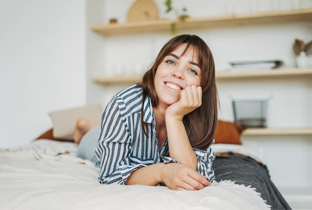 Young happy brunette woman in casual clothes rest on bed at the bright interior