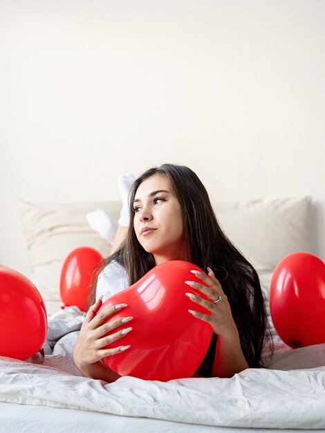 Young happy brunette woman laying in the bed with red heart shaped balloons