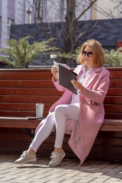 A young happy business girl in sunglasses is studying a folder with documents sitting on a wooden bench on the street in the city Next to a notebook and a thermocup of coffee Vertical photo