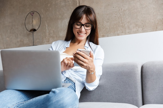 young happy business woman indoors at home on sofa in living room using laptop computer using mobile phone.