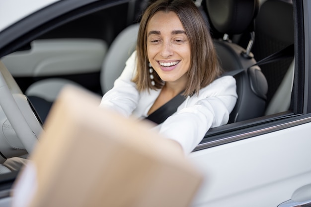 Photo young and happy business woman receiving a parcel through the vehicle window on the go while driving a car. concept of fast delivery on the go