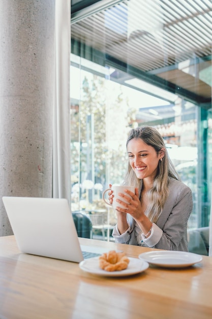 Young happy business woman using laptop, drinking tea or coffee from a mug in modern open workplace