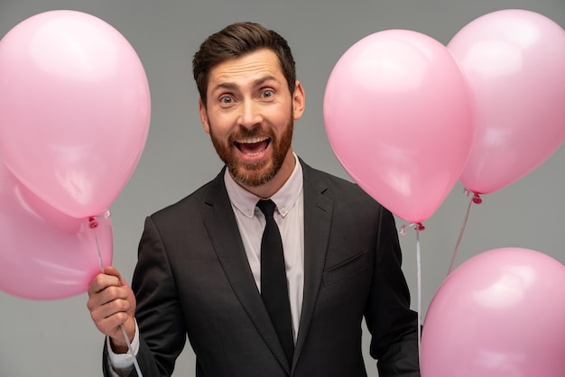 Young happy businessman wearing formal suit looking through a lot of pink balloons to camera