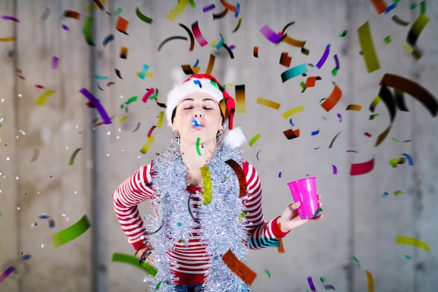 Young happy casual business woman wearing a red hat and blowing\
party whistle while having new years confetti party in front of\
concrete wall