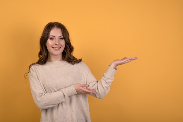 A young and happy caucasian brunette girl with wavy hair in a\
casual jumper pointing with her hand to a palm with an empty\
advertising space on an orange studio background.