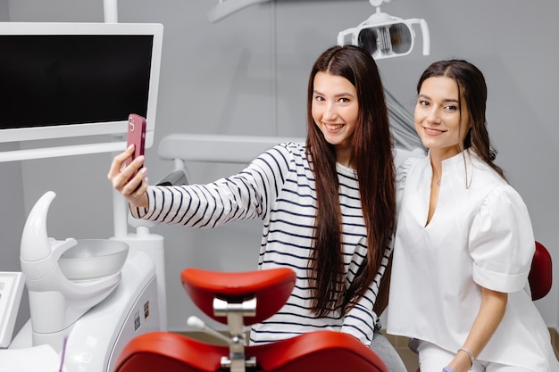 Young happy caucasian girl doing selfie shot on mobile phone sitting at dental office chair with female dentist