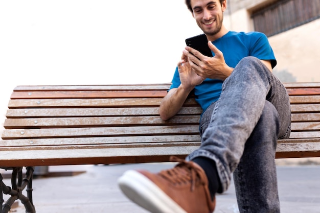 Молодой счастливый кавказский человек расслабляющий сидя на скамейке с помощью мобильного телефона. Выборочный фокус под рукой. Скопируйте пространство. Социальные сети, технологии и концепции образа жизни.
