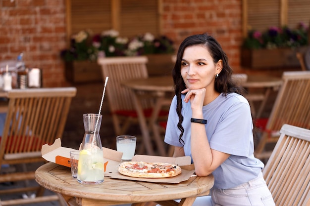 Young happy caucasian woman sitting at table eating freshly baked pizza and drinking fresh in outdoor cafe on clear sunny summer day