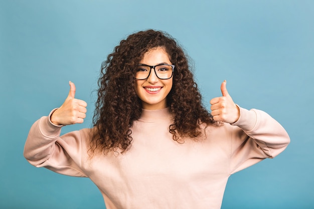 Young happy cheerful curly woman showing thumb up isolated over blue background.