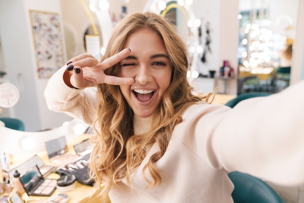 Foto la giovane ragazza bionda allegra felice all'interno nel salone di bellezza prende un selfie che mostra il gesto di pace.