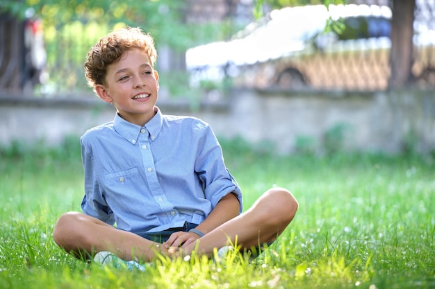 Young happy child boy relaxing on green grass in summer park. Positive kid enjoying summertime outdoors. Child wellbeing concept.