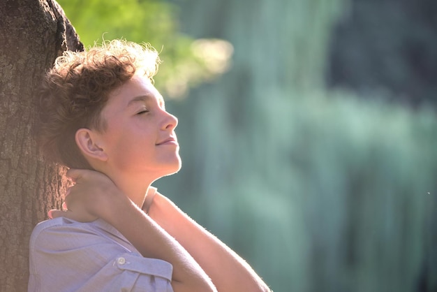 Молодой счастливый мальчик отдыхает в летнем парке, опираясь на ствол дерева Концепция благополучия ребенка