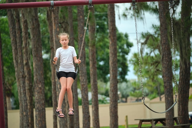 Young happy child girl playing alone flying high on swings on\
summer weekend sunny day safety and recreation on playground\
concept