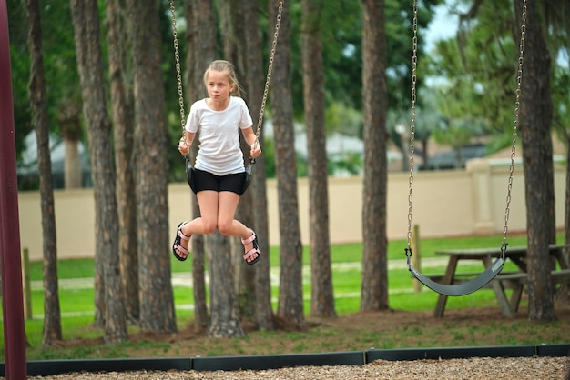 Young happy child girl playing alone flying high on swings on summer weekend sunny day Safety and recreation on playground concept