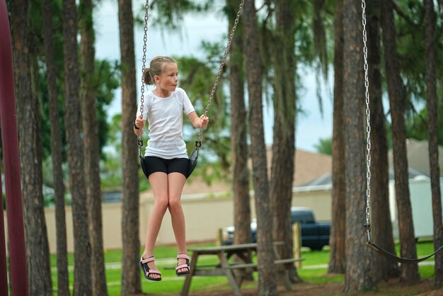 Young happy child girl playing alone flying high on swings on summer weekend sunny day Safety and recreation on playground concept