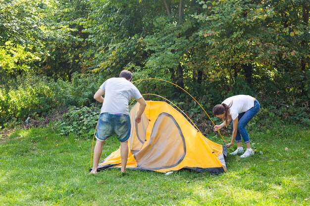 Young happy couple are preparing for camping. They're installing a tent on a suitable place in a meadow.