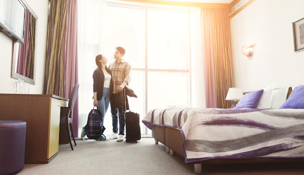 Young happy couple arrived to hotel room on honeymoon. Family standing on window background