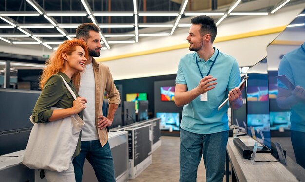 Young happy couple came to store of household appliances and\
gadgets to buy new plasma tv. sales consultant helps young couple\
choose tv by showing showcase.