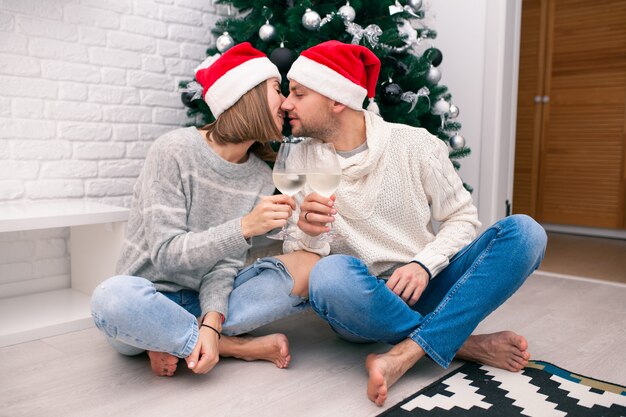 Young happy couple in Christmas hats near a Christmas tree kissing, holding glasses of wine. New Year celebration