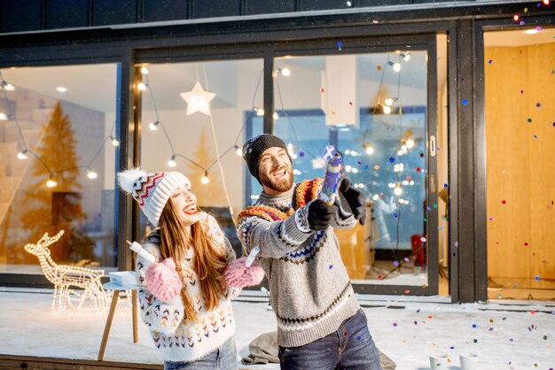 Young happy couple dressed in sweaters celebrating winter holidays in front of a beautiful decorated house in the mountains