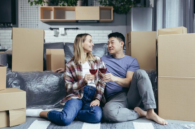 Young happy couple drinking wine in glasses sitting on the floor in a new apartment Asian man and woman happy together among cardboard boxes just moved