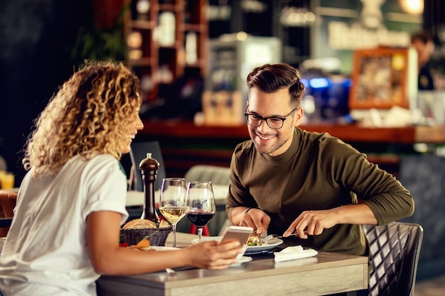 Giovane coppia felice che mangia in un bar e legge qualcosa sullo smartphone focus è sull'uomo