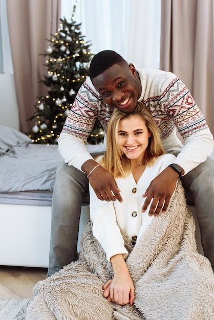 Young happy couple embracing and relaxing on comfortable bed. Lovely multiethnic couple wearing christmas sweater and hugging. African man and affectionate woman near christmas tree.