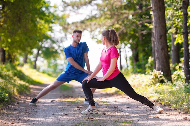 young happy couple enjoying in a healthy lifestyle warming up and stretching before jogging on a country road through the beautiful sunny forest, exercise and fitness concept