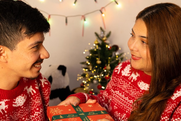 Photo young happy couple give each other gifts wearing christmas sweatshirts with christmas lights