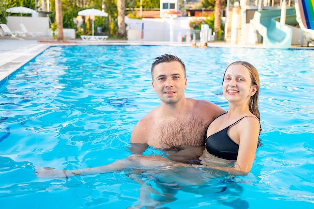 A young happy couple a guy and a girl are swimming in the pool on a water slide having fun on vacation smiling and laughing