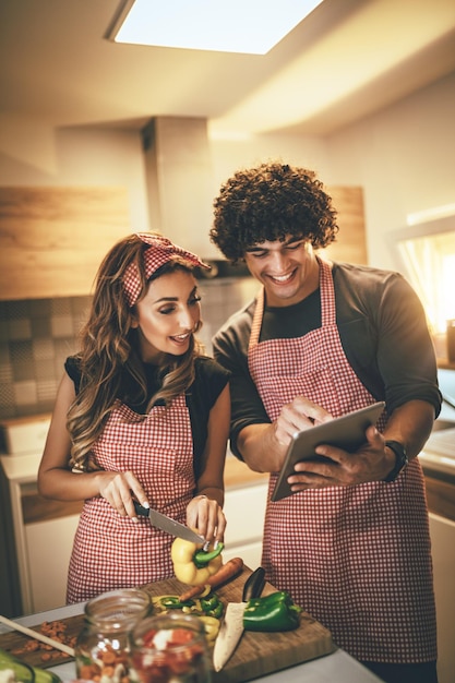 Photo young happy couple is enjoying and preparing healthy meal in their kitchen and reading recipes on the tablet.