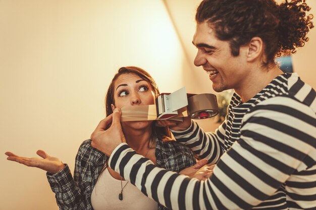 The young happy couple is moving into a new house. They are packing boxes and having fun. Young man bonding adhesive tape on a mouth his girlfriend and playing with her.