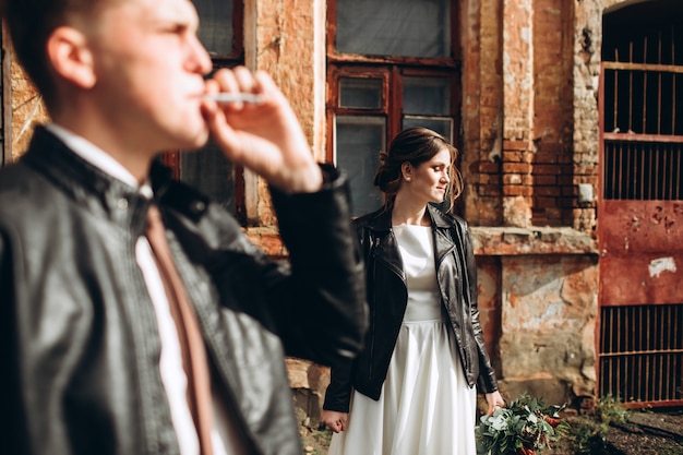 Young happy couple is walking on a city street after rain. Bride and groom with leather jackets outdoors. Autumn wedding.