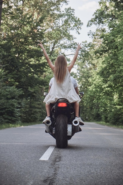 A young happy couple rides a motorcycle on an asphalt road in\
the forest freedom and speed backside