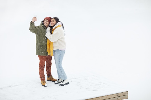 Молодая счастливая пара улыбается в камеру, делая селфи-портрет на мобильном телефоне во время прогулки зимой