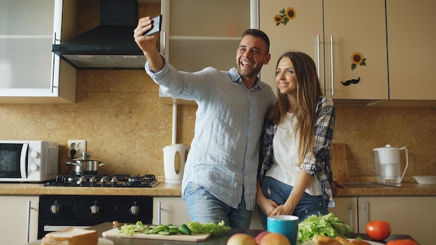 Young happy couple taking selfie picture while cooking breakfast in the kitchen at home