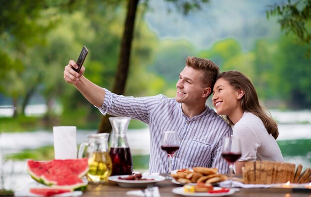 young happy couple taking selfie while having picnic french dinner party outdoor during summer holiday vacation  near the river at beautiful nature