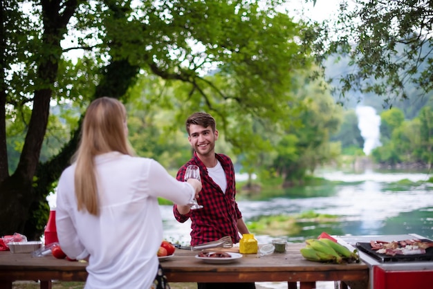 young happy couple toasting red wine glass while having picnic french dinner party outdoor during summer holiday vacation  near the river at beautiful nature