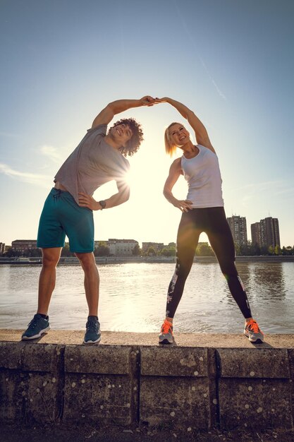 Young happy couple training outdoors by the river, stretching together on the wall at sunset.